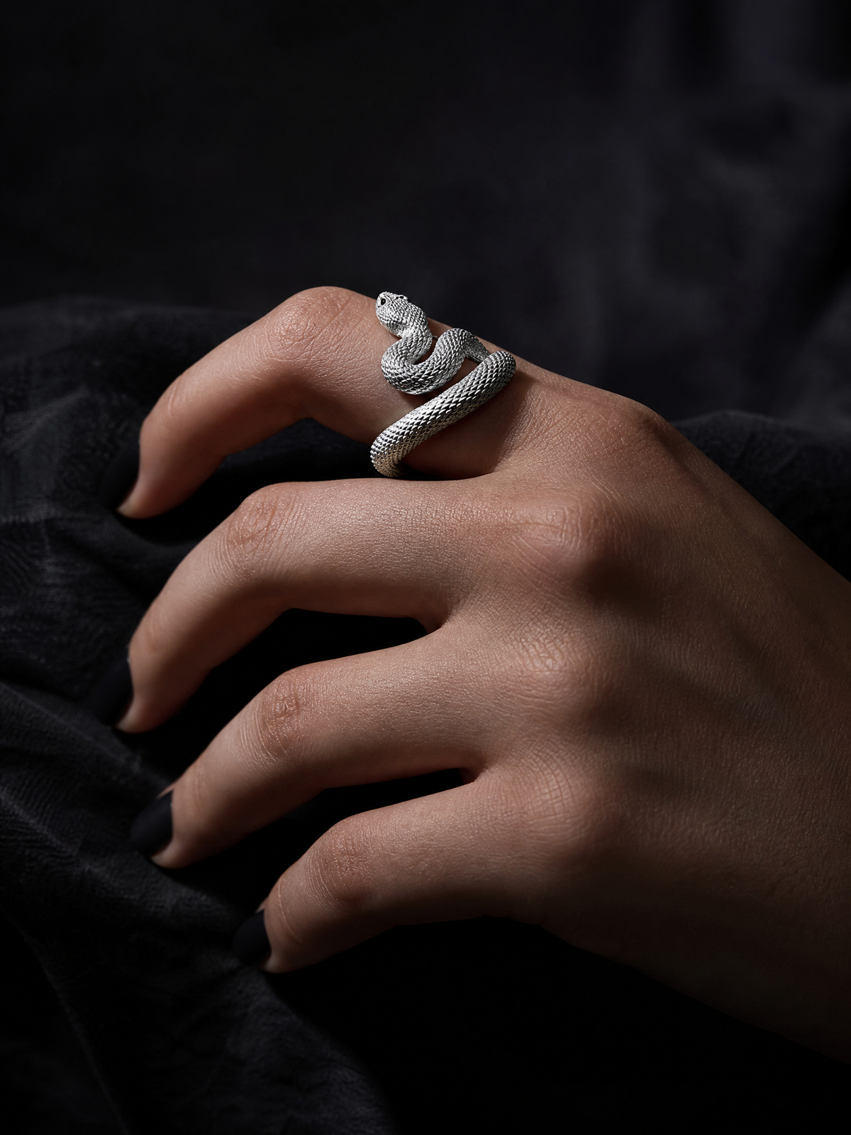 Serpent's Coil silver rattlesnake ring worn on a hand with black nails against dark fabric